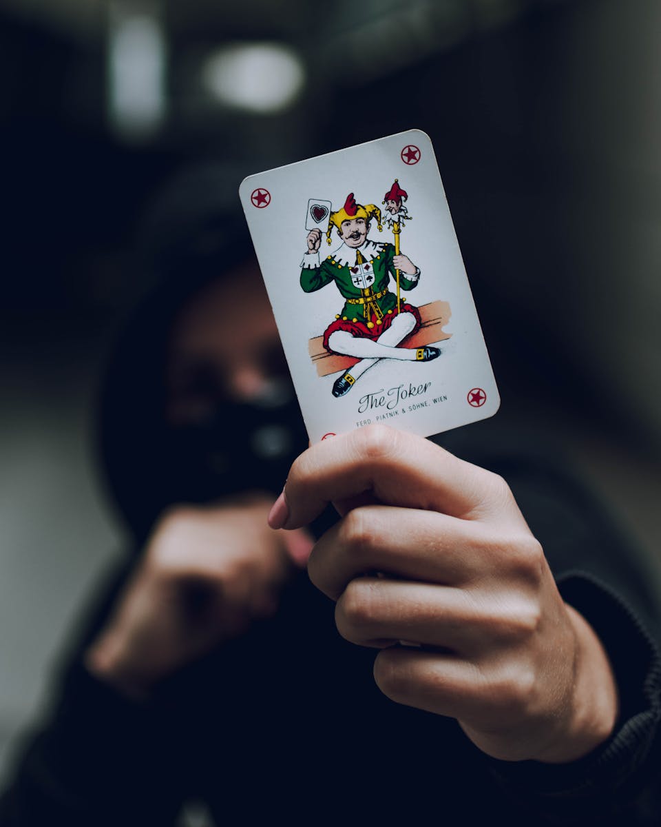 Mysterious hooded figure holding a joker card in a dimly lit setting, emphasizing intrigue and drama.